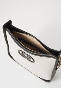 Sac en toile avec des finitions noires et un logo bien visible. L'intérieur est doublé de beige et comporte des compartiments zippés.