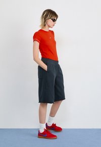 Rød figursyet t-shirt med hvidt logo, mørkegrå shorts med sidelommer og røde sneakers med blå accenter. Model står mod en hvid baggrund.