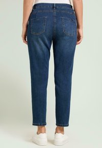 Pantalones vaqueros de mezclilla azul oscuro con un diseño ajustado y entallado. Incluyen dos bolsillos traseros y una cinturilla. Combinados con calzado blanco.