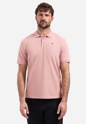 Man met kort bruin haar, gekleed in een lichtroze poloshirt en zwarte broek, staand tegen een effen lichte achtergrond.