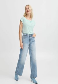 Hellblaues kurzärmeliges Oberteil mit gerafftem Ausschnitt, kombiniert mit weit geschnittenen, ausgeblasten blauen Jeans und spitzen blauen Absätzen. Einfaches und sauberes Design.