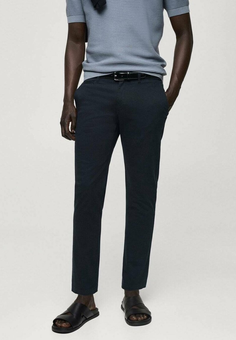 Homme portant une chemise en maille bleu clair à manches courtes, ceinture noire, pantalon slim bleu marine et sandales noires à bout ouvert, debout les mains dans les poches.