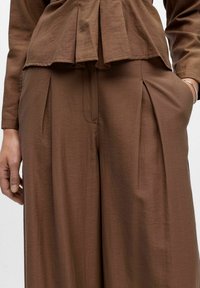 Gros plan d'une personne portant un pantalon ample marron plissé et un haut assorti, avec une main dans la poche et des bracelets en argent au poignet.