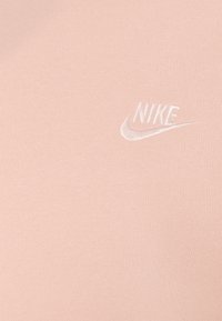 Světlé růžové plátno s malým bílým vyšívaným logem Nike a swoosh na horní pravé straně.