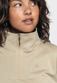 Beige, hochkragiger Pullover mit einem halben Reißverschluss, glatter Textur und einem kleinen gestickten Logo auf der Brust. Hat einen taillierten Schnitt.