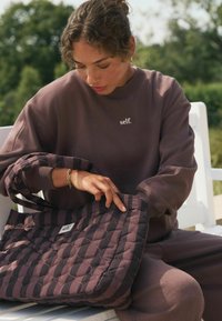 Geantă tote matlasată în nuanțe de mov închis cu o textură modelată. Modelul poartă un hanorac supradimensionat asortat cu logo-ul "self.".