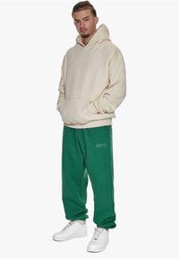 Beige Kapuzenpullover mit Fronttasche und geprägtem Logo, kombiniert mit grünen Sweatpants mit Seitentaschen und Logo, getragen mit weißen Sneakers.