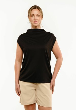 Kobieta o jasnych włosach, ubrana w czarną bezrękawnikową bluzkę i beżowe spodenki, stojąca na tle jednolitego białego tła, zwrócona przodem.