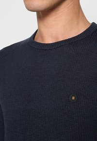 Marineblauwe gebreide trui met een textuurstof, geribde ronde halslijn en een klein zeshoekig zwart-goud logo op de borst.