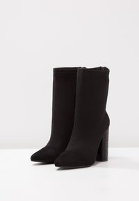 Botines de ante negro con un tacón cuadrado texturizado, punta afilada y un diseño de tobillo ajustado y suave. Sin cierres visibles.