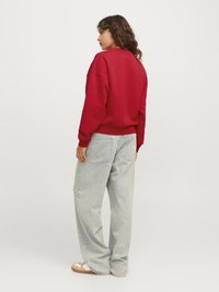 Felpa rossa oversize con spalle scese, abbinata a jeans a gamba larga blu chiaro e slip-on color beige. La texture è liscia; il design è casual.