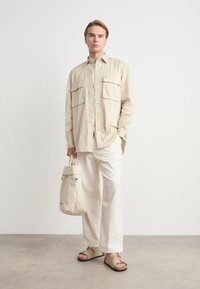 Beige overhemdd met twee voorzakken, gecombineerd met witte wijde broek en beige sandalen. Model houdt een beige tote bag vast.