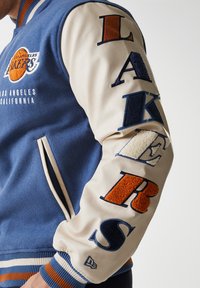 Blue wool varsity jacket with cream leather sleeves, featuring "LAKERS" in multi-colored raised letters. Includes logo and striped cuffs.
