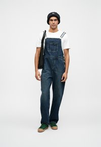Dunkelblaue Denim-Latzhose mit verstellbaren Trägern, Fronttasche und lockerer Passform. Kombiniert mit einem weißen T-Shirt und grünen Sneakers.
