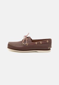 Braune Lederbootsschuh mit beiger Gummisohle, ausgestattet mit goldenen Ösen, klassischer Schnürung und sichtbaren Nähten entlang der oberen Kante.