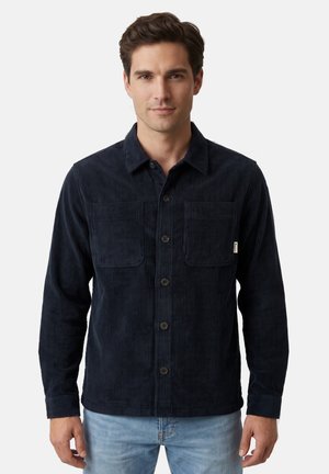 Hombre con una camisa de pana azul oscuro con botones y dos bolsillos en el pecho, y vaqueros azul claro, de pie frente a un fondo liso.