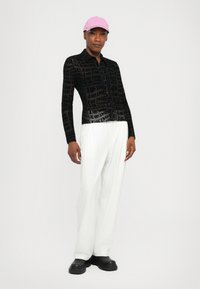 Black sheer button-up shirt with repeated "JEANS" text, white striped pants, and a pink cap. Chunky black shoes complete the look.