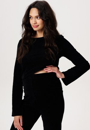 Schwarzes Cord-Top mit langen Ärmeln, das einen Ausschnitt in der Taille aufweist. Kombiniert mit passenden schwarzen Cord-Hosen, beide besitzen eine weiche Textur.