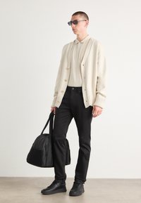 Krämfärgad cardigan med runda knappar, beige pikétröja, svarta jeans och svarta sportskor. Håller en svart sportväska och bär mörka solglasögon.