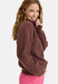 Suéter marrón de punto con cuello alto, con un patrón de diamantes y textura acanalada. Combinado con pantalones de pana rosa.