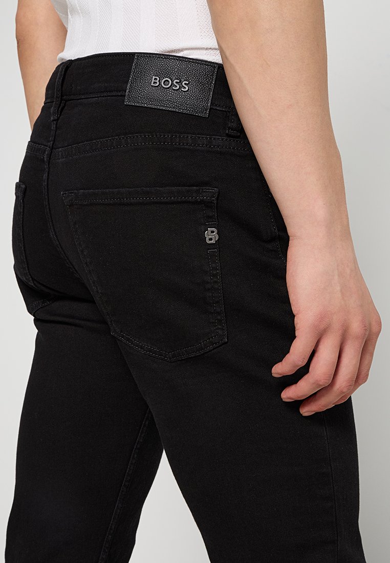 Gros plan sur une personne portant un jean noir BOSS, montrant la poche arrière, le patch en cuir BOSS, et la main droite reposant sur le côté.
