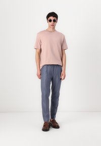 Helles rosa Kurzarm-T-Shirt, lockere Passform; blaue maßgeschneiderte Hose; braune Lederschuhe; runde Sonnenbrille mit dünnem Rahmen.