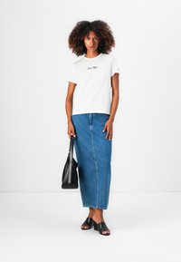 T-shirt blanc à manches courtes avec texte brodé, associé à une longue jupe en denim bleu. Des sandales à talons noires et un sac à bandoulière noir complètent le look.