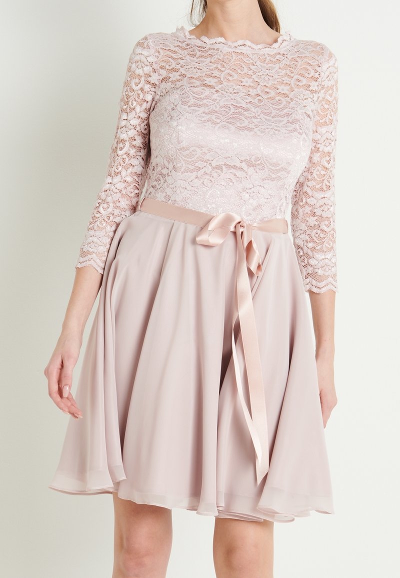 Femme portant une robe rose pâle avec des manches longues en dentelle et un ruban en satin noué à la taille, jupe flottant au-dessus des genoux.
