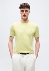 Νέος άνδρας όρθιος με τα χέρια στις τσέπες, φοράει ανοιχτό κίτρινο t-shirt και λευκό παντελόνι με μαύρη ζώνη, ουδέτερο φωτεινό φόντο.