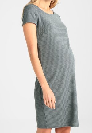 Vrouw draagt een grijze zwangerschapsjurk met korte mouwen en knielengte met subtiele horizontale strepen, staand tegen een effen lichte achtergrond.