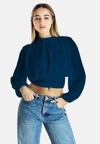 Blusa blu cropped con maniche a sbuffo e colletto alto, abbinata a jeans in denim a vita alta con lavaggio chiaro. Tessuto dalla superficie liscia.