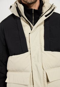Chaqueta acolchada con una sección superior negra y una sección inferior crema, con un cuello alto, cierre delantero con cremallera y grandes bolsillos frontales.