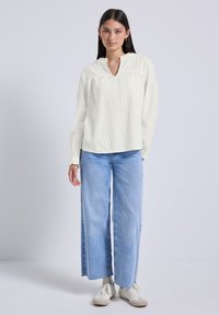Donna con lunghi capelli scuri che indossa una blusa bianca a maniche lunghe con texture, jeans a gamba larga blu chiaro e sneakers bianche, in piedi contro uno sfondo semplice.