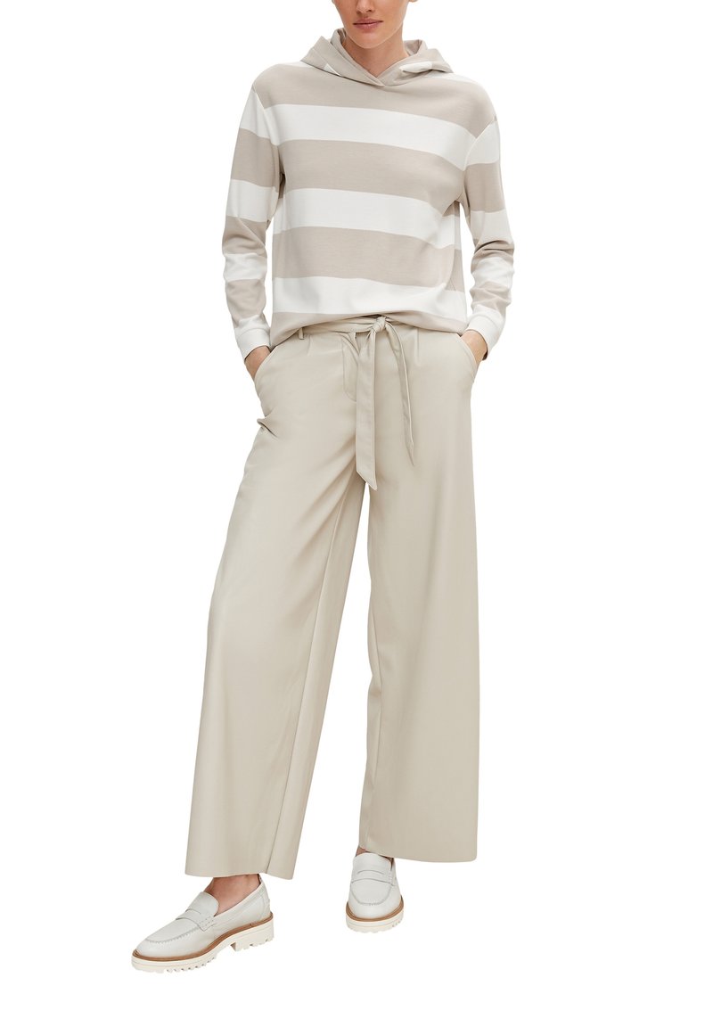 Femme portant un sweat à capuche beige et blanc à larges rayures, pantalon beige à jambes larges noué à la taille, mocassins blancs, debout les mains dans les poches.