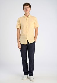 Korte mouwen, overhemd met knopen in lichtgele stof met een borstzak, gecombineerd met donkerblauwe broek en witte sneakers.