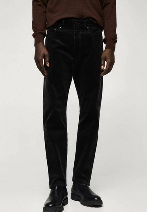 Man draagt zwarte corduroy broek, zwarte leren laarzen en een donkerbruin shirt met lange mouwen, staand tegen een effen achtergrond.