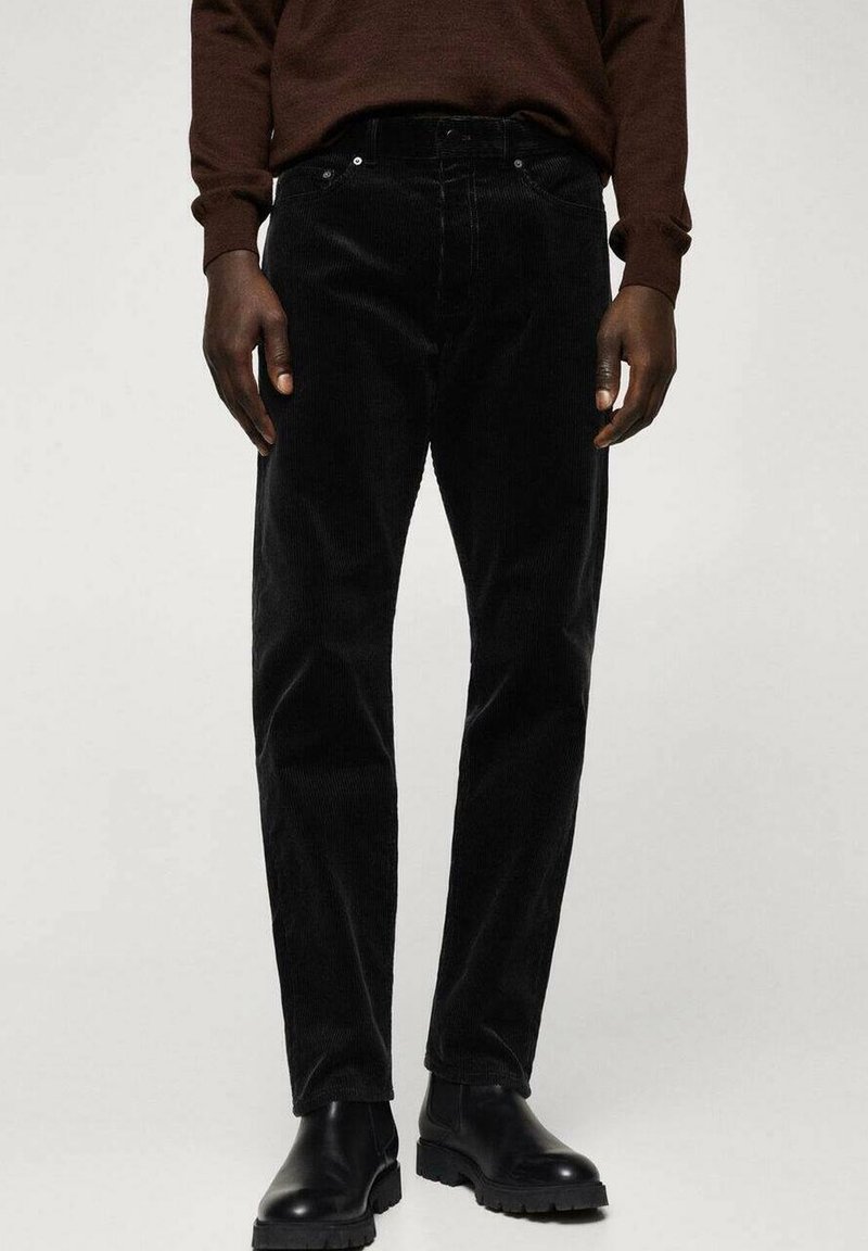 Homme portant un pantalon en velours côtelé noir, des bottes en cuir noir et une chemise à manches longues marron foncé, debout devant un arrière-plan uni.