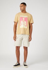 T-shirt in cotone beige chiaro con stampa grafica in rosa e rosso, abbinata a pantaloni cargo chiari. Sneakers in bianco e nero completano l'outfit.