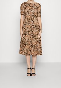 Brun paisley-mönstrad klänning med båtformad krage och korta ärmar, med en utsvängd kjol. Ett par svarta klackar med ankelsnörning.