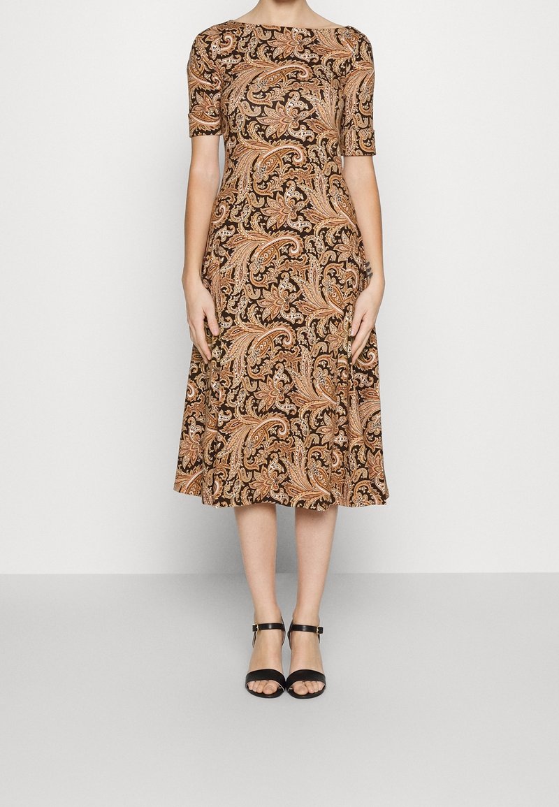 Brun paisley-mönstrad klänning med båtformad krage och korta ärmar, med en utsvängd kjol. Ett par svarta klackar med ankelsnörning.