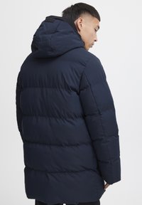 Puffjakke i marineblå, med hette, ribbestrikkede mansjetter og quiltet tekstur. Designet vektlegger varme og en avslappet passform.