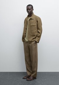 Brown linen jacket with front pockets, olive green t-shirt, brown linen trousers, and black sandals on a figure against a neutral background.