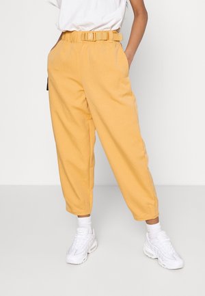 Persoon die een hoog getailleerde gele broek met riem draagt, witte sokken, witte chunky sneakers en een ingestopt wit T-shirt tegen een effen achtergrond.