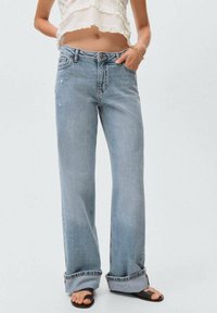 Jean en denim bleu clair à taille haute, à jambes larges, avec des détails déchirés et des ourlets roulés, associé à un top court blanc.