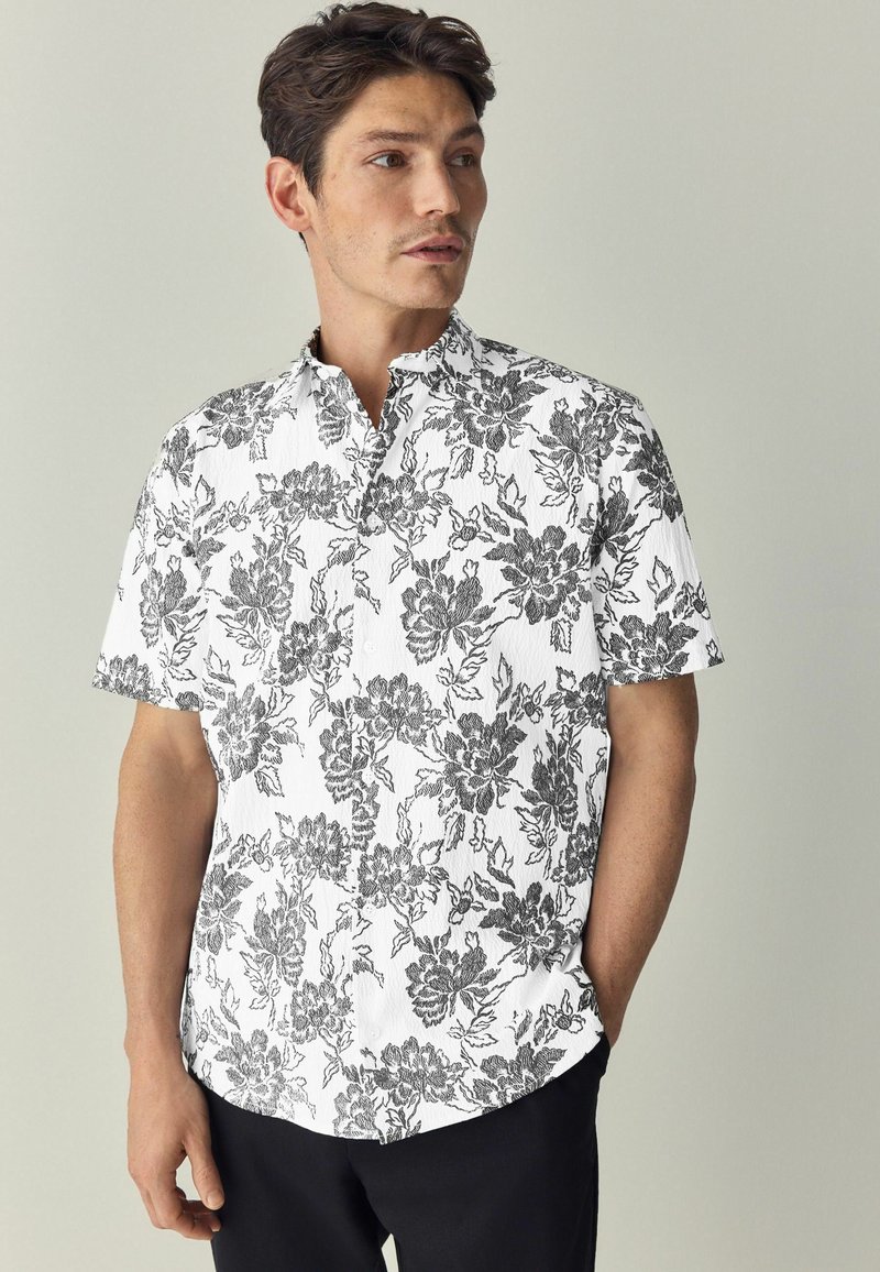 Camicia a maniche corte bianca con un motivo floreale nero. Presenta un colletto rialzato e un tessuto testurizzato. Abbinata a pantaloni scuri.