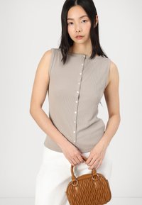 Top sans manches beige à côtes avec un col rond et un détail de boutons sur le devant, associé à un sac à main texturé marron et un pantalon blanc.