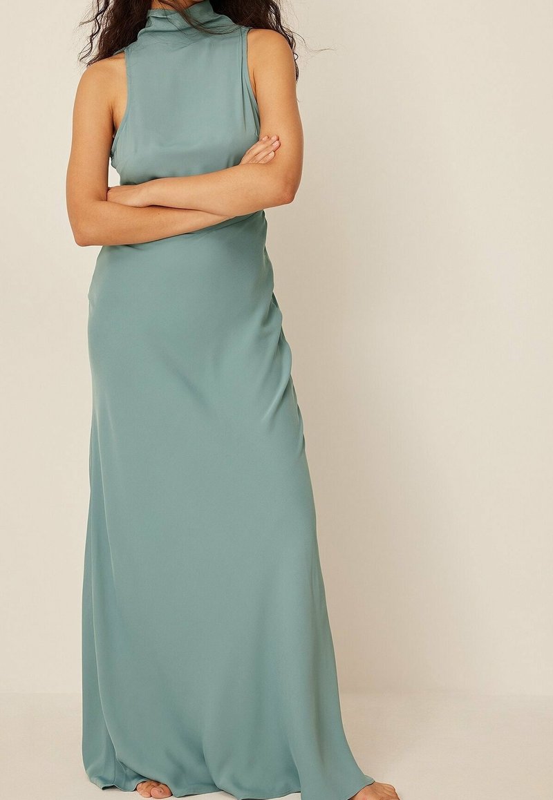 Femme debout, les bras croisés, portant une robe sans manches, longue jusqu'au sol, de couleur bleu-vert clair, sur un fond beige uni.