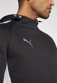 Zwarte sportieve hoodie met grijs Puma-logo, heeft een hoge kraag, trekkoord met plastic hardware en witte zijkanten. Gladde textuur.