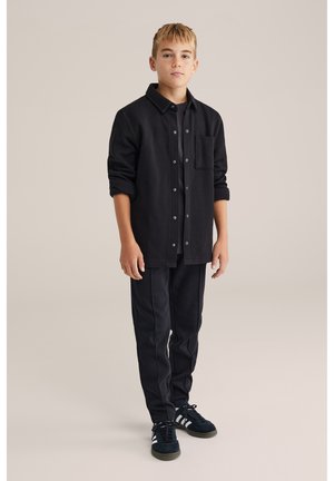 Melnā krekla jaka ar teksturētu virsmu un plāksteru kabatu, kombinēta ar melnām šaurām biksēm. Melni sporta apavi ar baltiem akcentiem.