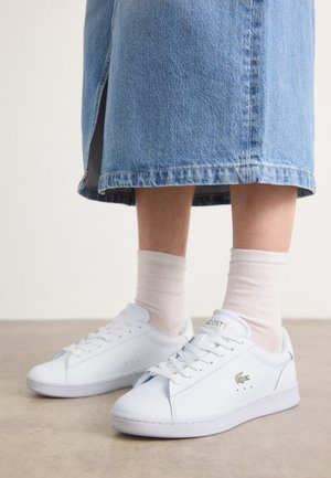 Person, der bærer hvide sneakers med Lacoste-logo, lysebeige sokker og en blå denimbukser med sideslids, stående på et glat gulv.